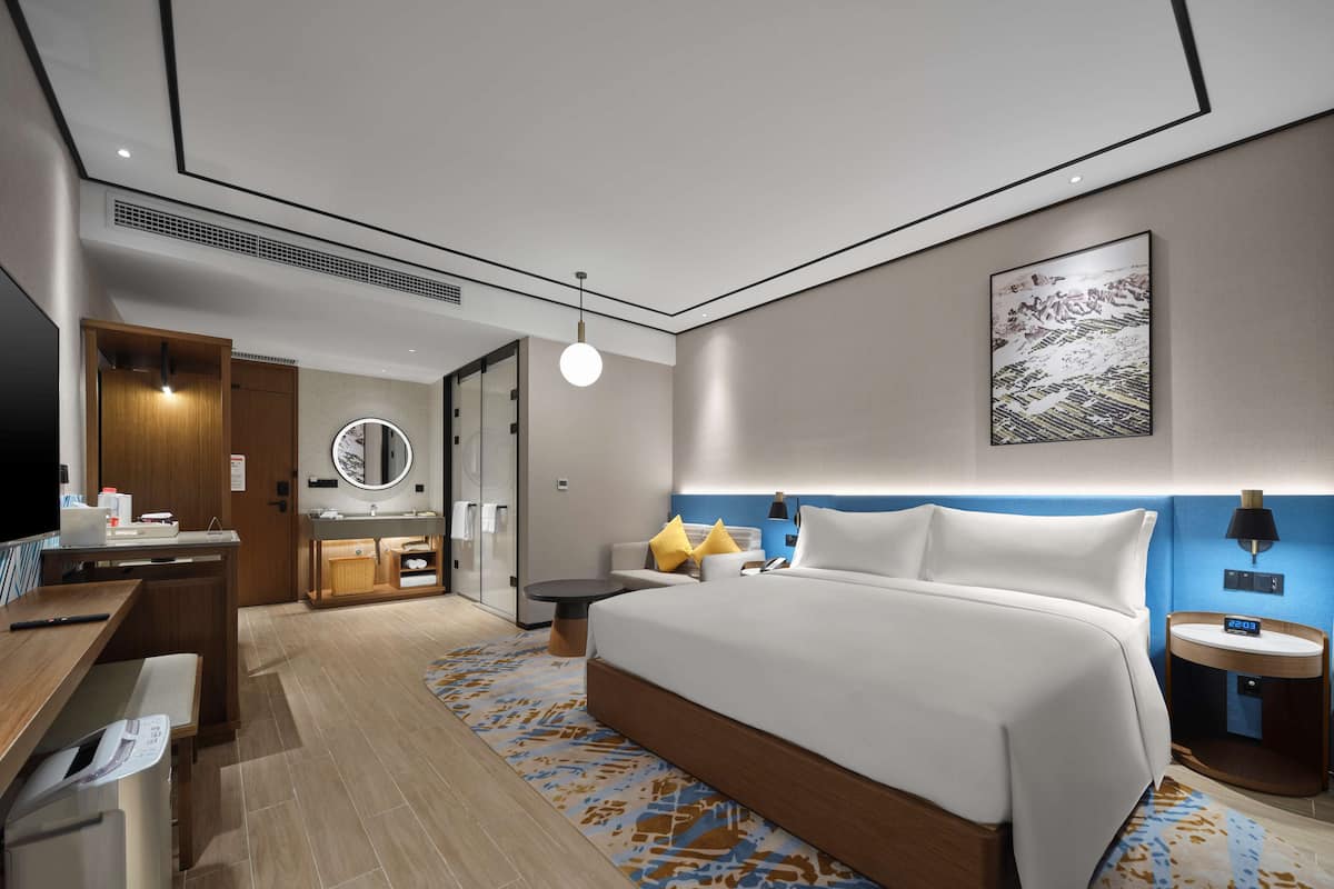 Ein modernes Hotelzimmer mit einem großen Bett, einem Schreibtisch mit Computer, einem Badezimmer mit Spiegel und einem Wandbild.