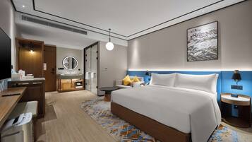 A modern hotel room with a large bed, a desk with a computer, a bathroom with a mirror, and a wall-mounted artwork.