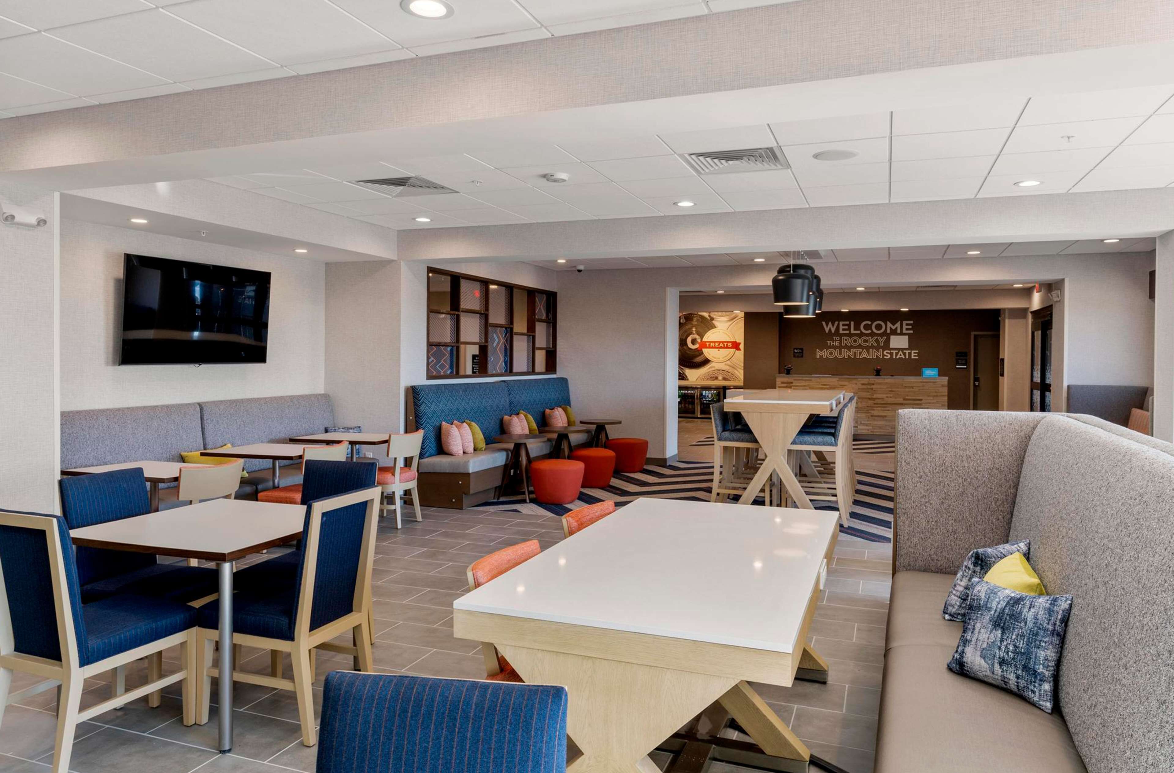 A modern hotel lobby with seating areas, a TV mounted on the wall, and a 'Welcome' sign.