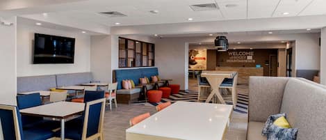 A modern hotel lobby with seating areas, a TV mounted on the wall, and a 'Welcome' sign.