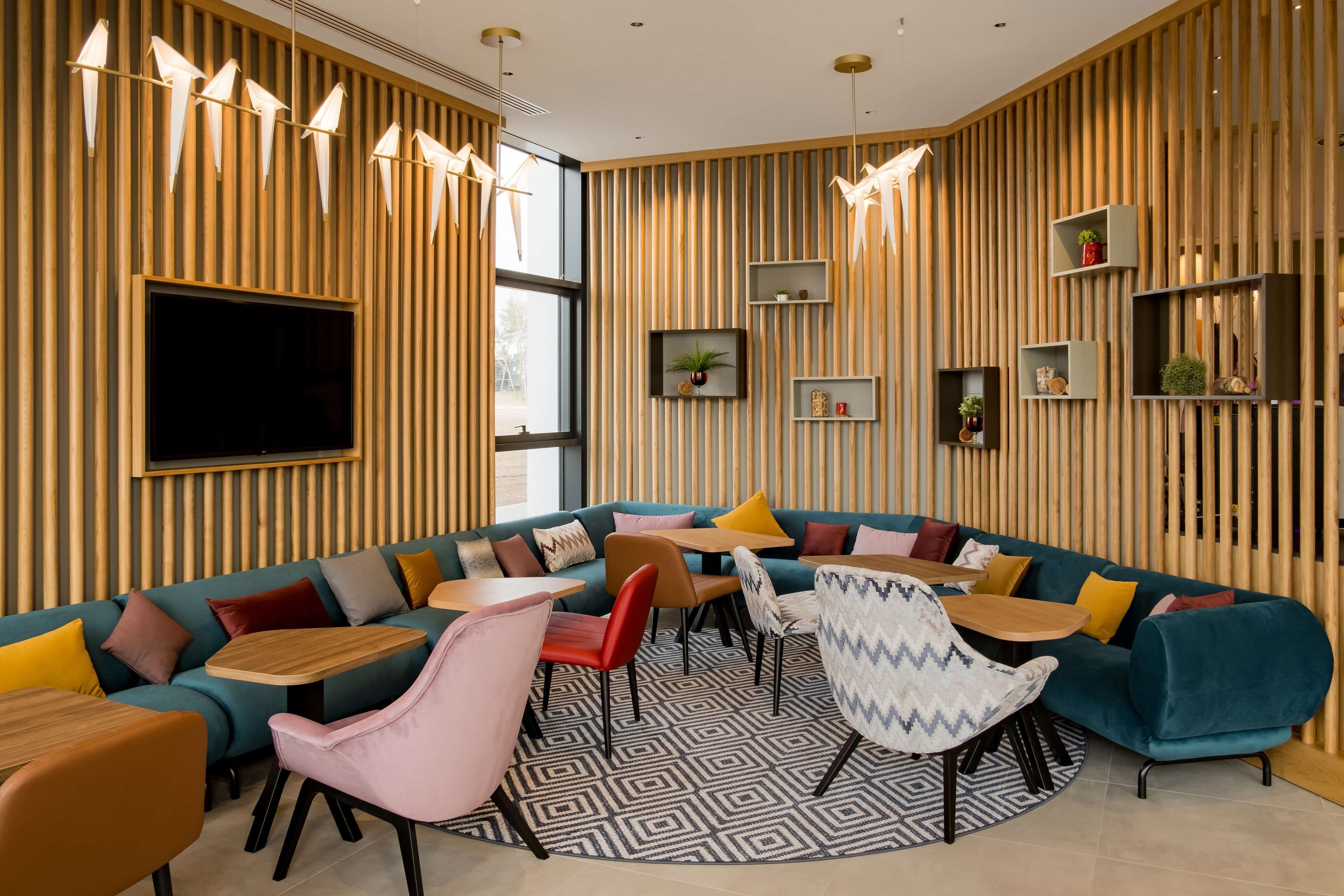 A modern lounge area with a curved sofa, multiple chairs, and tables, wooden paneling on the walls, and a flat-screen TV mounted above the sofa.