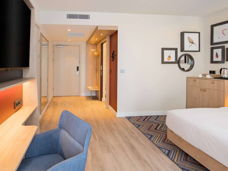 Een hotelkamer met een bed, een bureau, een stoel, een televisie, een kledingkast en een gang die naar een andere kamer leidt.