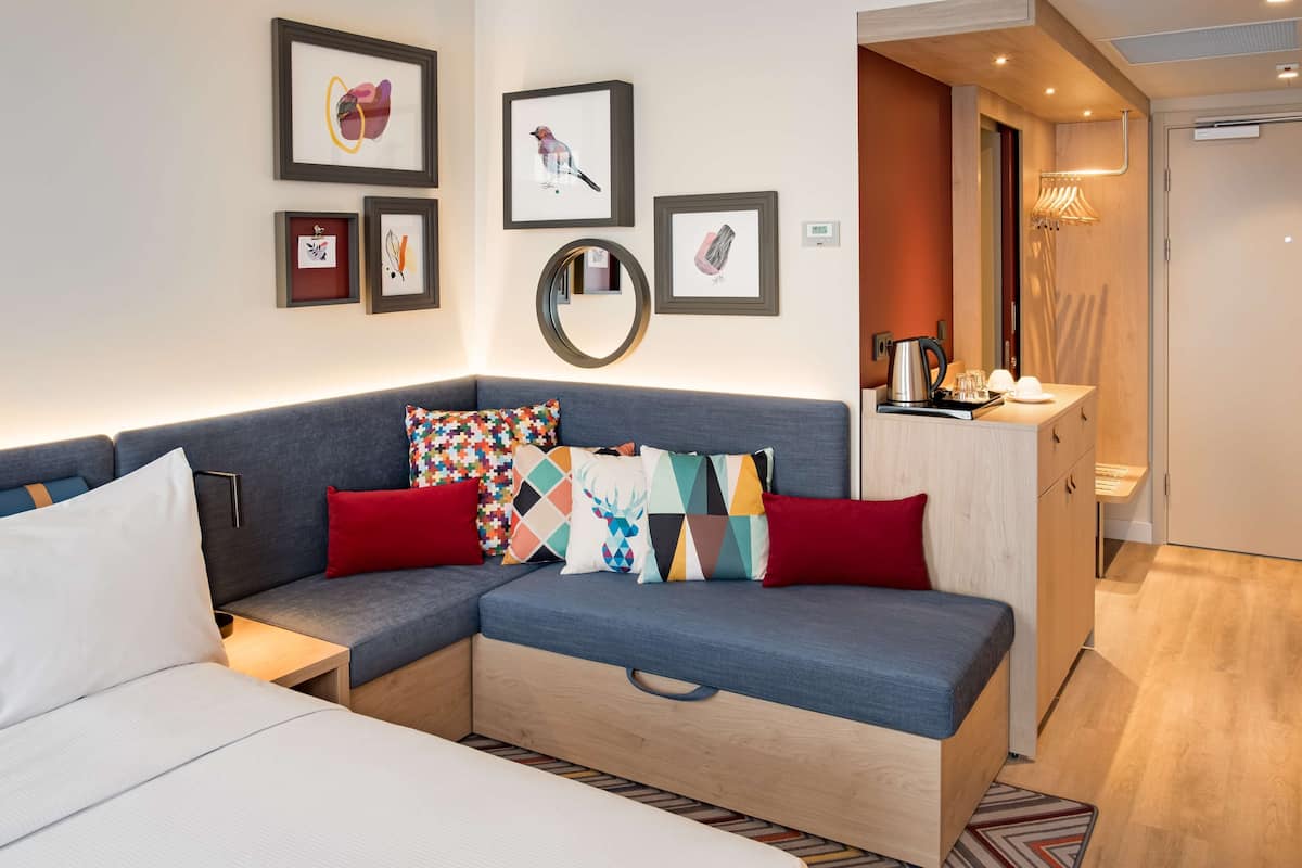 A modern hotel room with a grey sofa, colorful cushions, a small round mirror, and framed artwork on the wall.