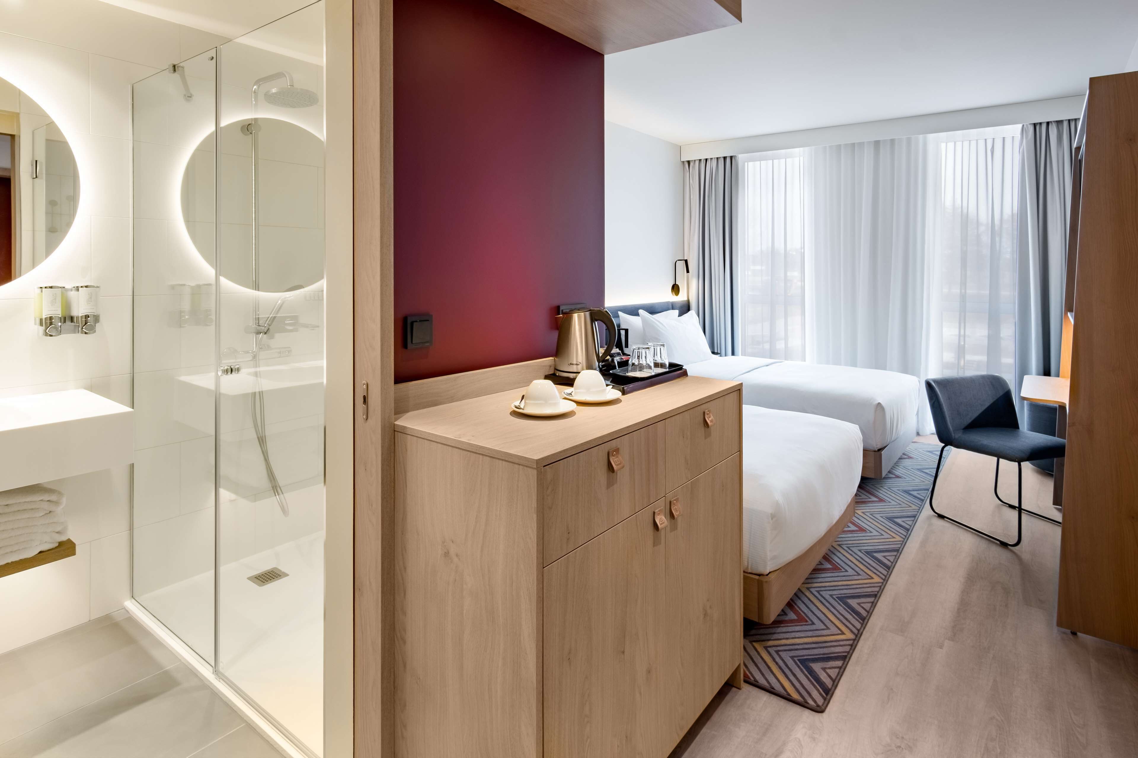 Hotellihuone, jossa on lasiseinällinen suihku, puinen lipasto, sänky, jossa on kaksi tyynyä, työpöytä tuolilla sekä suuri ikkuna, jossa on verhot.