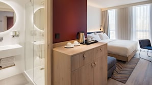 Ein modernes Hotelzimmer mit einer gläsernen Dusche, einem hölzernen Waschtisch mit Spiegel, einem Bett mit weißen Leinentüchern und Blick ins Freie durch große Fenster.