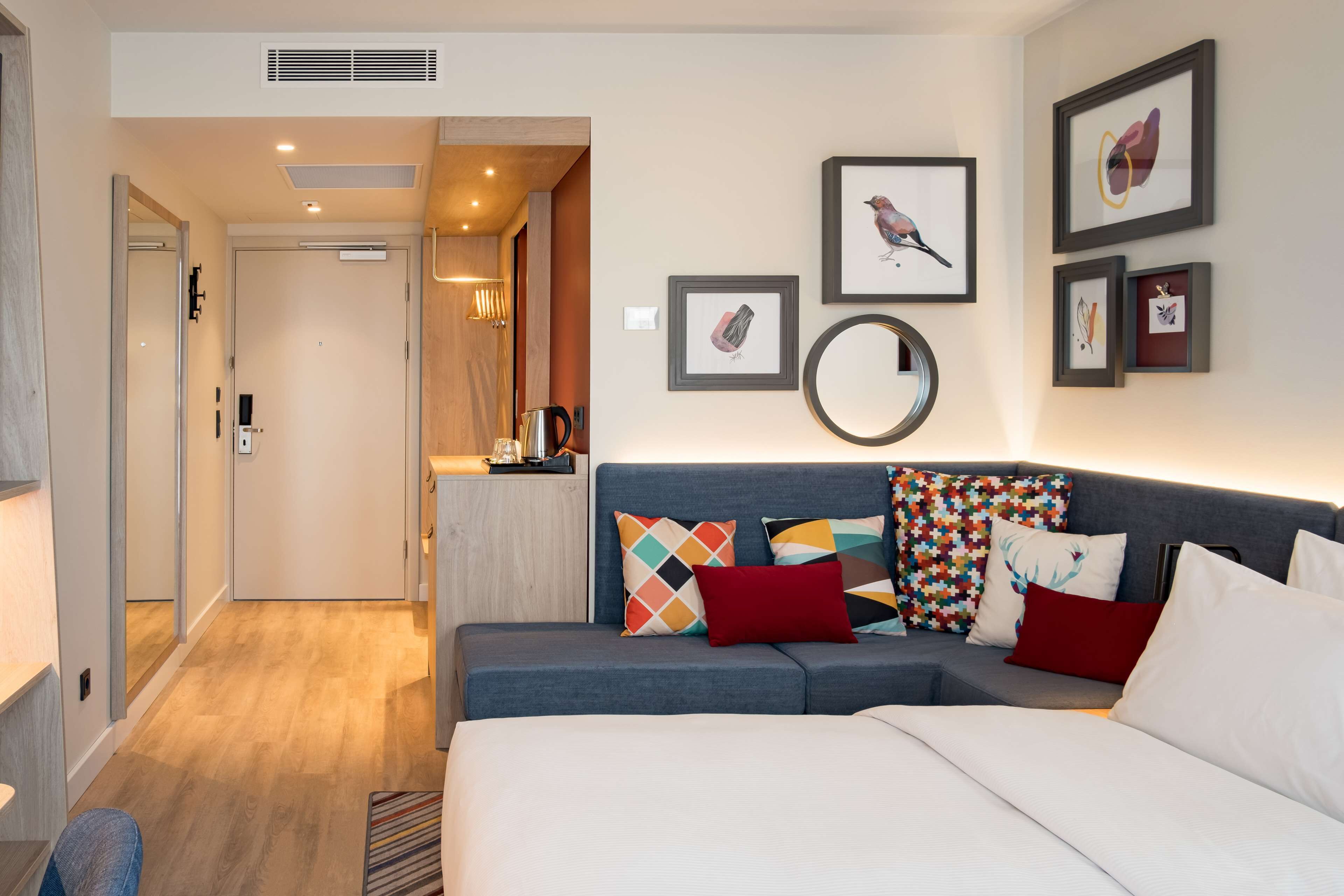 A modern hotel room with a large bed, a sofa, a desk, and a kitchenette. There are framed pictures on the wall and a mirror above the sofa.
