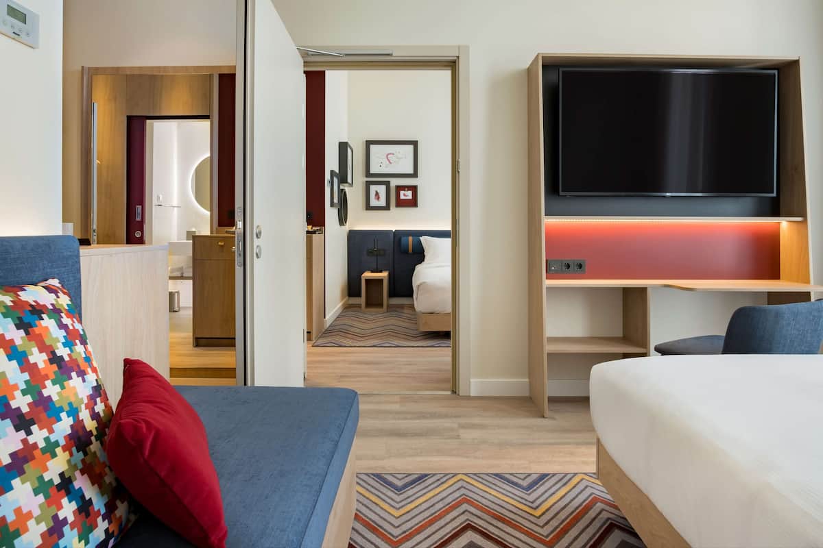 A hotel room with a bed, a desk with a TV, a bathroom, and a hallway leading to another room.