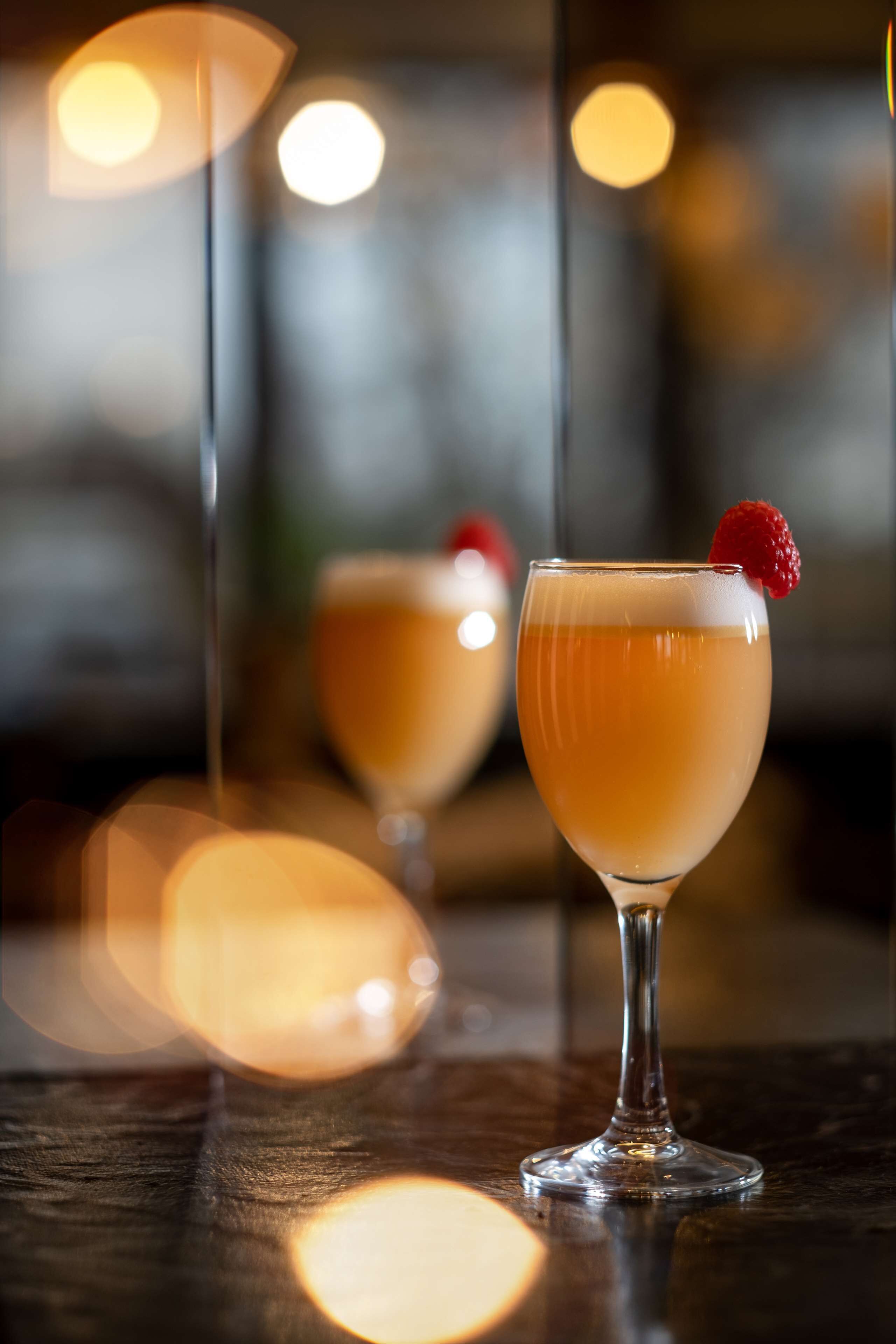 Deux cocktails servis dans des verres à pied, posés sur une surface en bois, avec des lumières floues en arrière-plan.