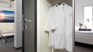 A modern bathroom with a glass shower enclosure, a white robe hanging on a rack, and a bed with a nightstand. - Sun Spalato Suite (Makarska)