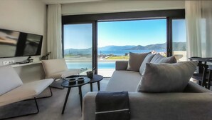A modern living room with a large window, a flat-screen TV, a sofa, a coffee table, and a view of the sea and mountains.