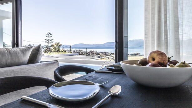 A dining area with a table set for two, a bowl of fruit, and a view of a coastal landscape.