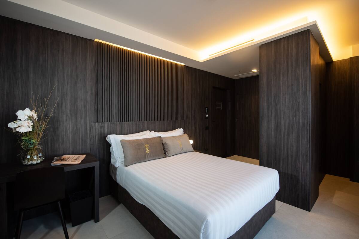 A modern bedroom with a large bed, dark wood paneling, a desk with a vase of flowers, and a nightstand.