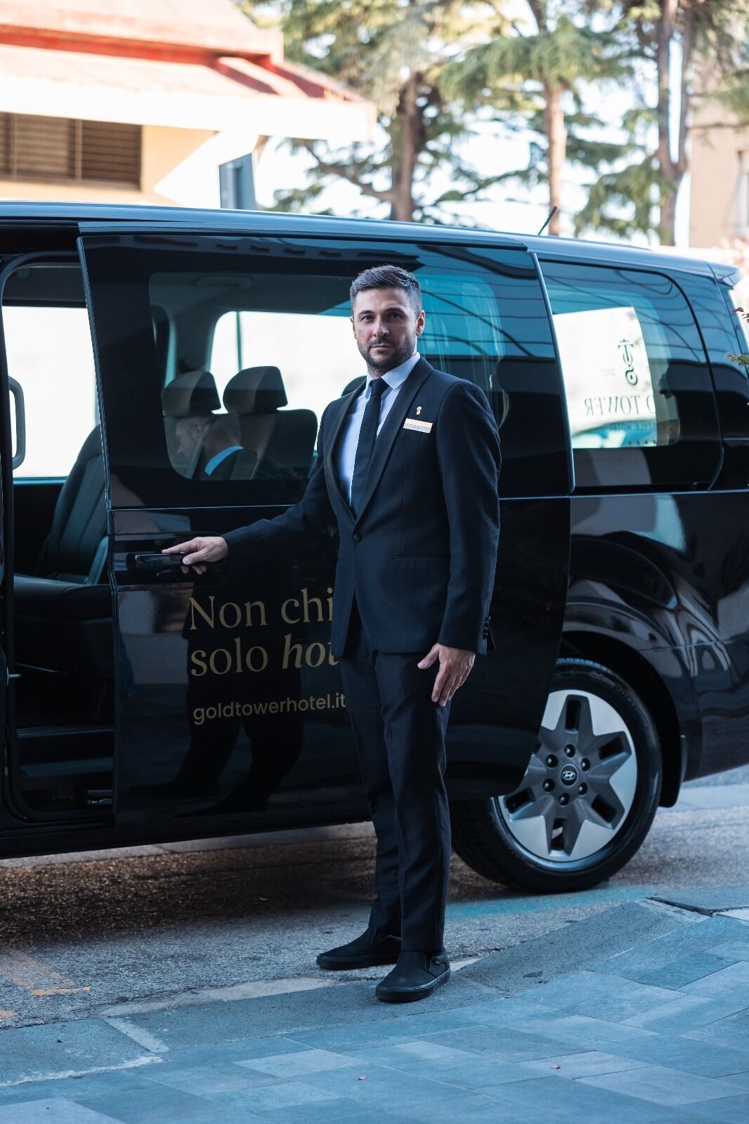 Ein Mann in einem Anzug steht neben einem schwarzen Van mit der Aufschrift 'Non chi solo hotel goldtowerhotel.it'.