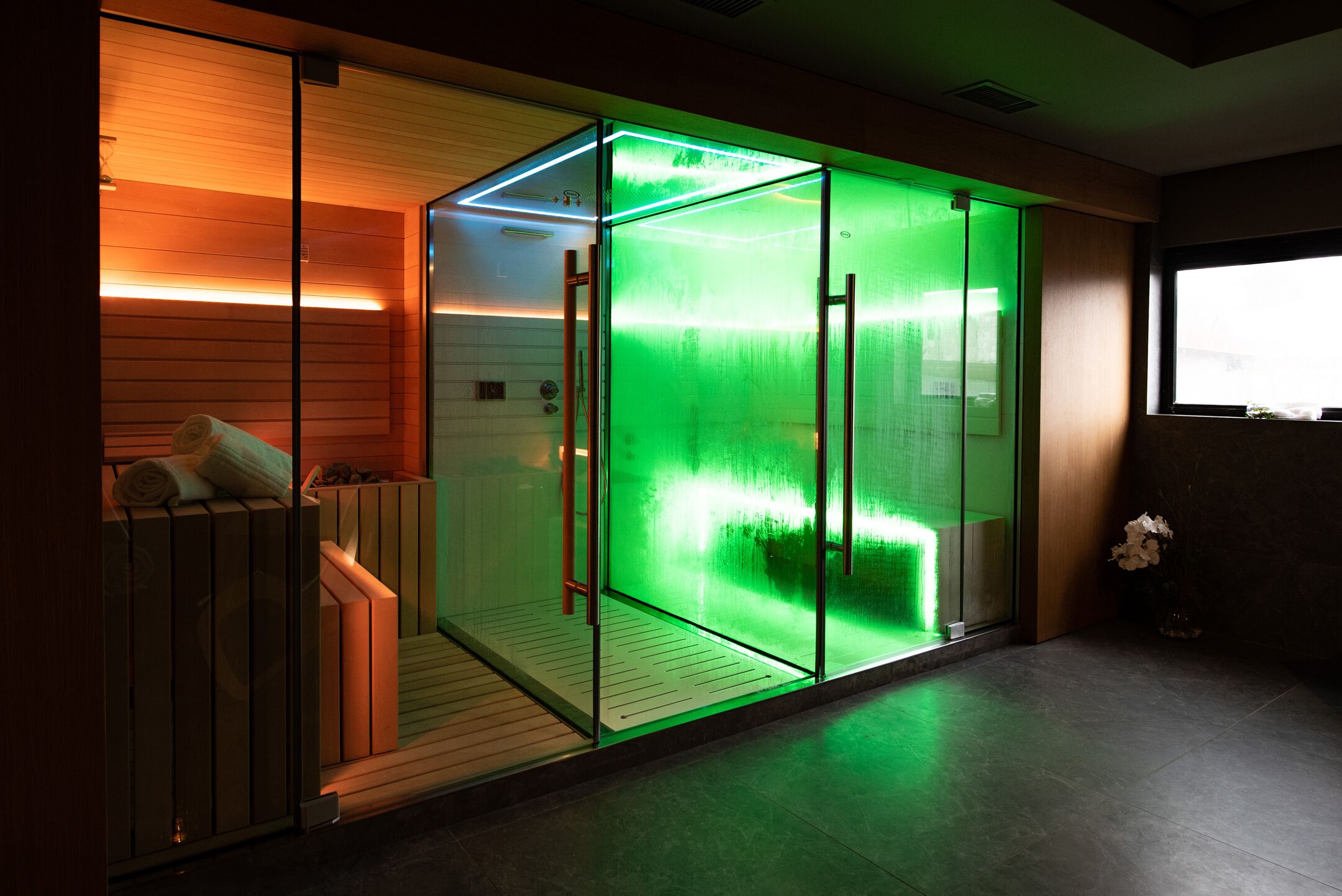 Una moderna spa con cabina in vetro, panche in legno e un elemento d'acqua illuminato di verde.