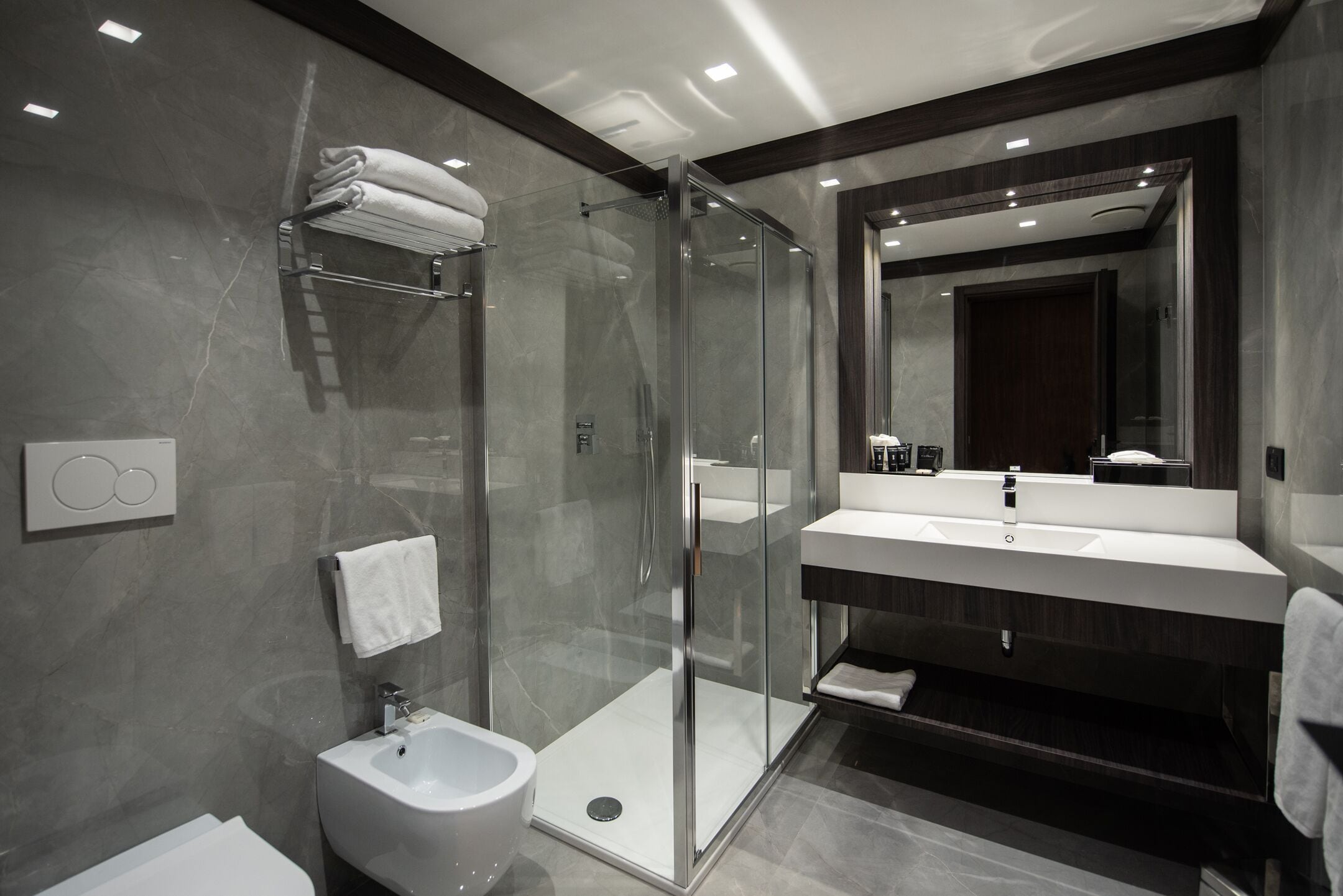 Ein modernes Badezimmer mit einer gläsernen Duschkabine, einem Bidet, einem Waschtisch mit Spiegel und einem Handtuchhalter.