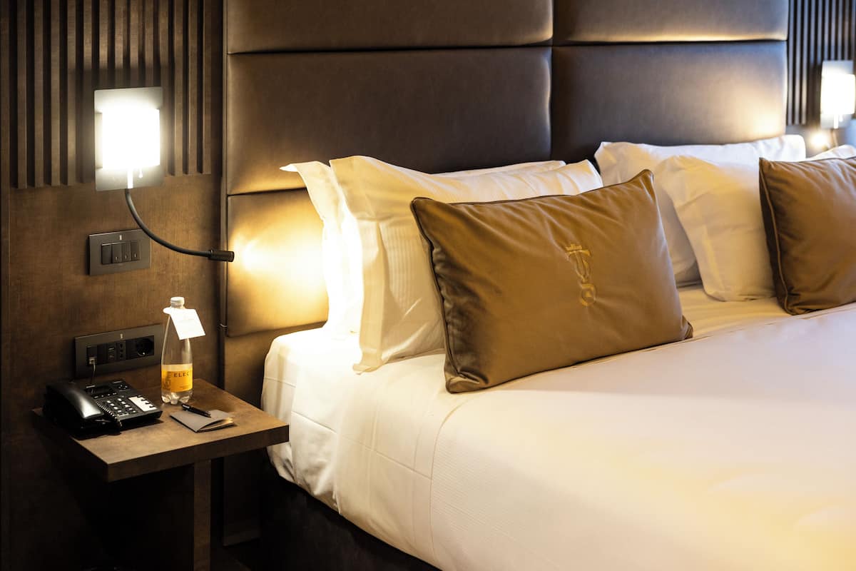 A hotel room with a bed, pillows, a bedside table with a phone and a bottle, and a wall-mounted light fixture.