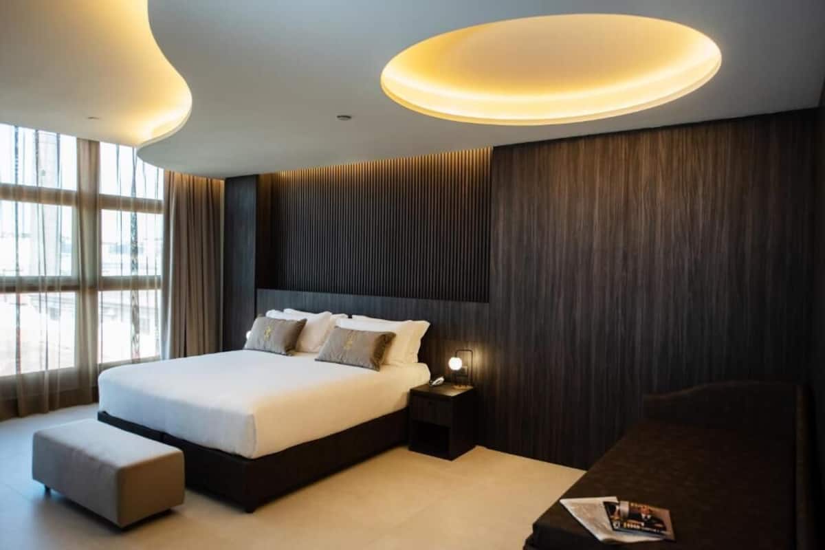 A modern hotel room with a large bed, a bedside table with a lamp, a small side table, and a view of the city through large windows.
