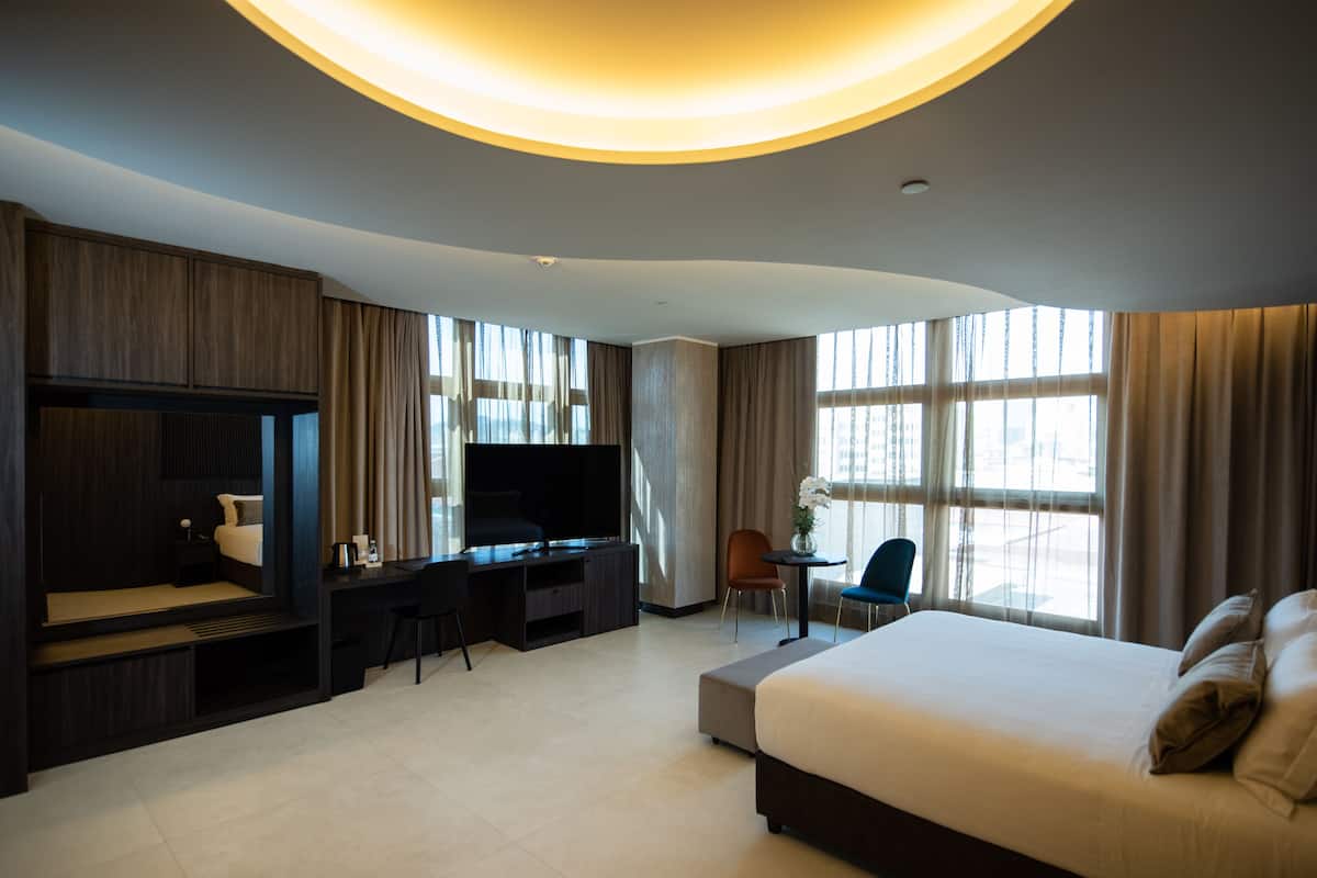 A modern hotel room with a large bed, a desk with a flat-screen TV, a chair, and a small table with a vase of flowers.
