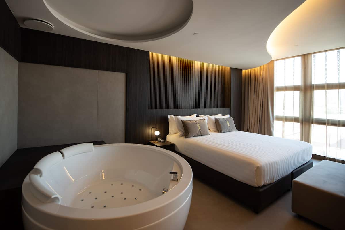A modern hotel room with a large bed, a built-in bathtub, and a large window providing natural light.