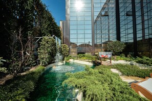 A modern building with a glass facade, a small outdoor pool with a waterfall, and a wooden bench. - Gold Tower Lifestyle Hotel (Naples)