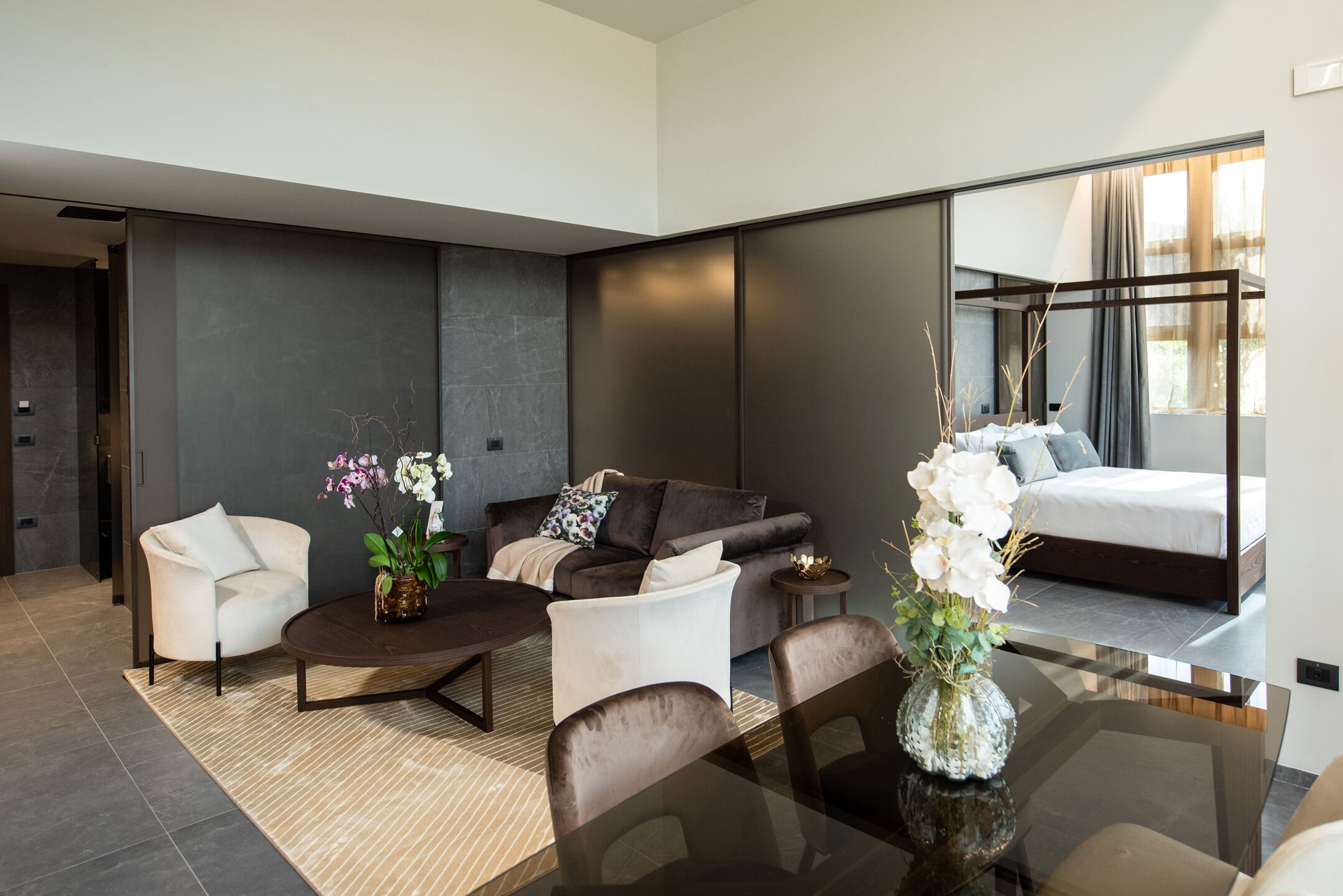 A modern hotel room with a sofa, armchairs, a coffee table, and a dining table. There is a large mirror reflecting the room and a bed with a view of the outdoors.