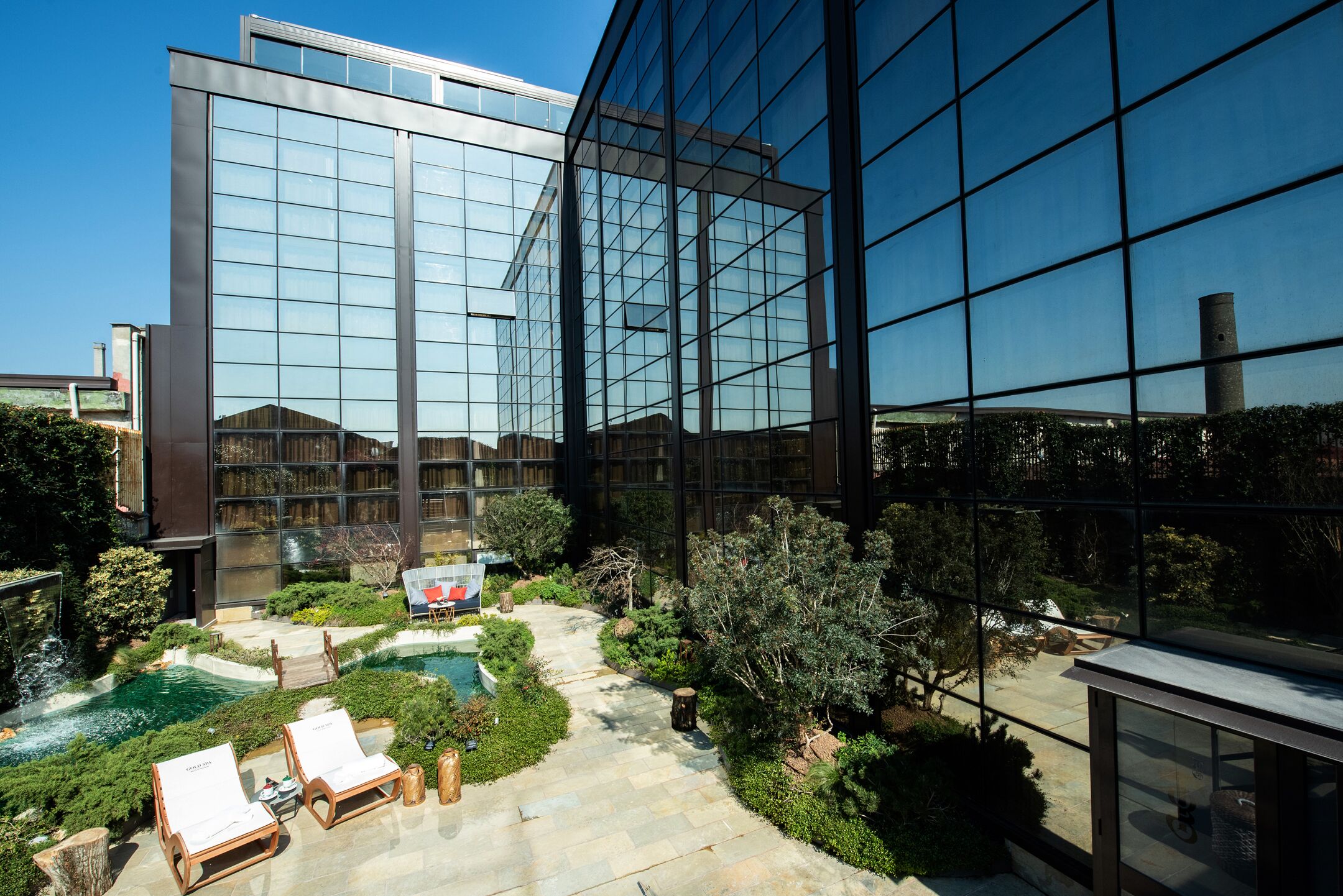 A modern building with a glass facade, a landscaped courtyard with a pool, and lounge chairs.