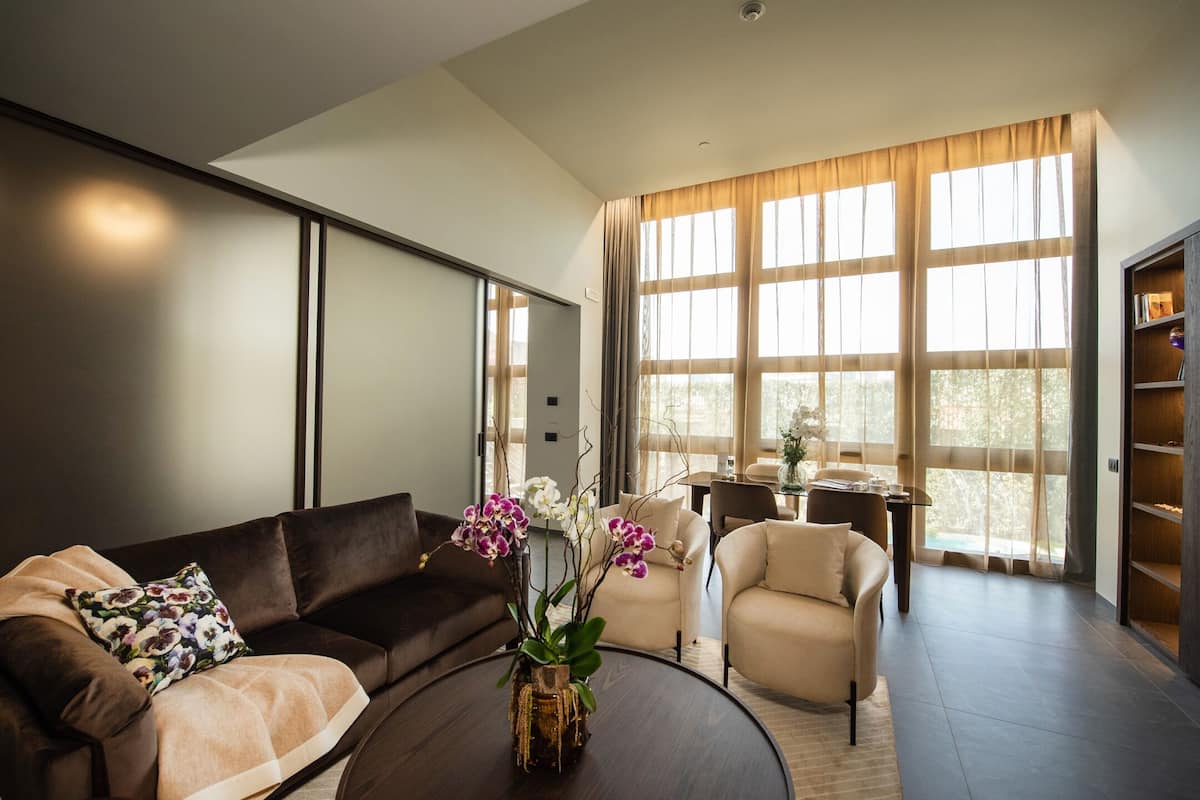 A modern living room with a dark brown sofa, a beige armchair, and a round coffee table with a vase of flowers. There is a large window with curtains, a desk with a chair, and a bookshelf.