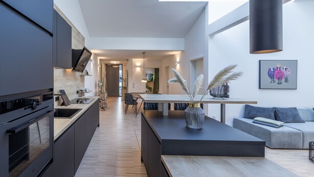 A modern kitchen with dark cabinetry, a central island, and a dining area with a sofa and table.