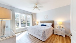 Une chambre avec un grand lit, deux tables de chevet avec des lampes, un ventilateur de plafond et une vue sur un immeuble par une grande fenêtre.