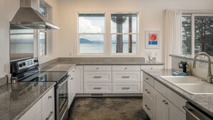A modern kitchen with granite countertops, white cabinetry, a stainless steel range hood, and a large window offering a view of the ocean. - Orcas Island, Peapod Cottages with Barn #290B (Orcas Island)