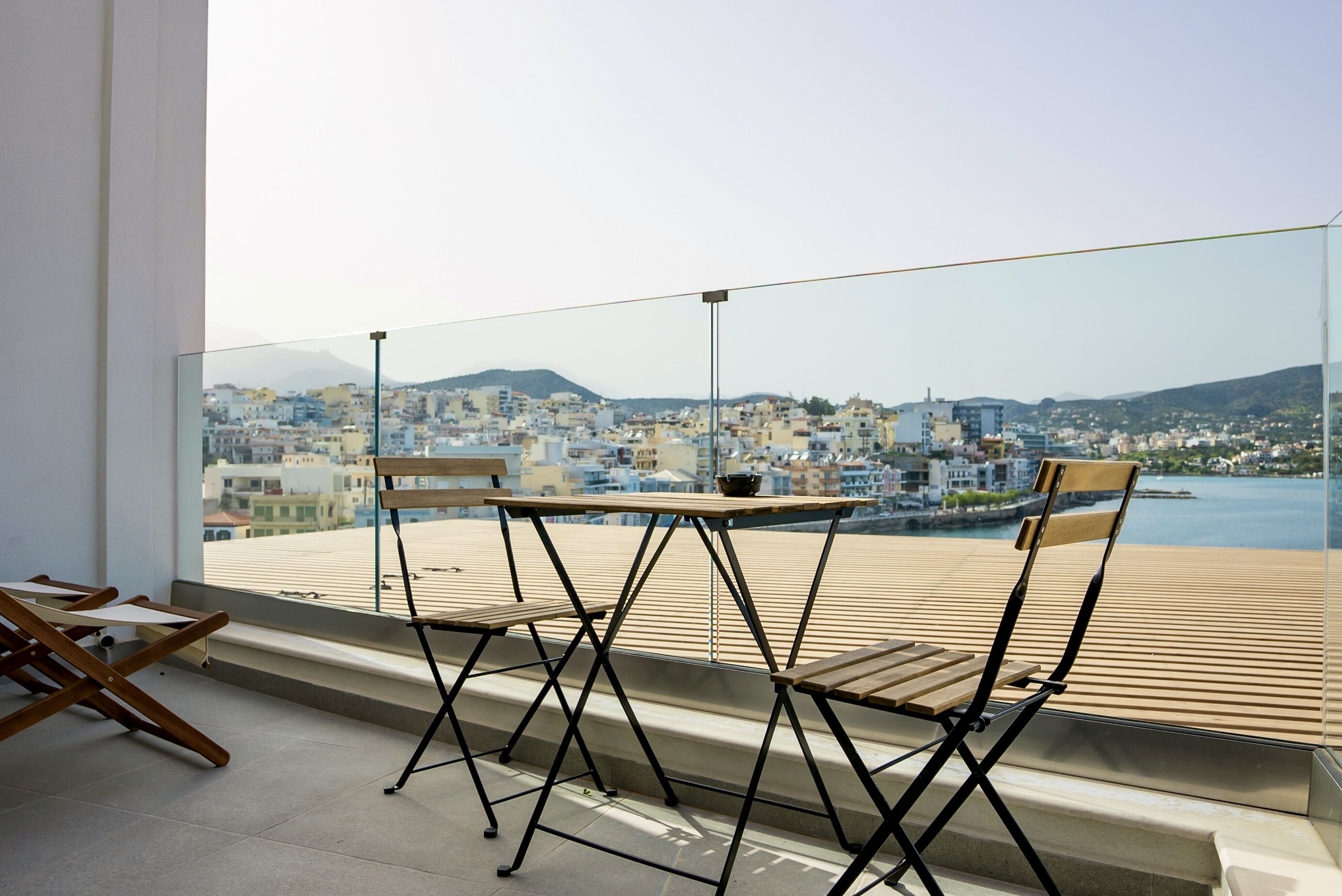 A balcony with two wooden chairs and a table, overlooking a cityscape with buildings and a body of water.