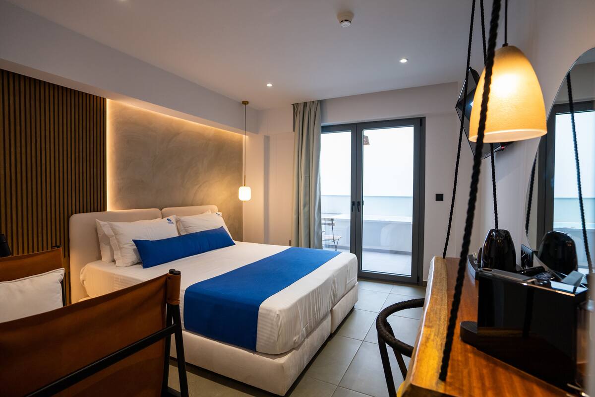 A hotel room with a bed, a desk, a chair, and a view of the sea through large windows.