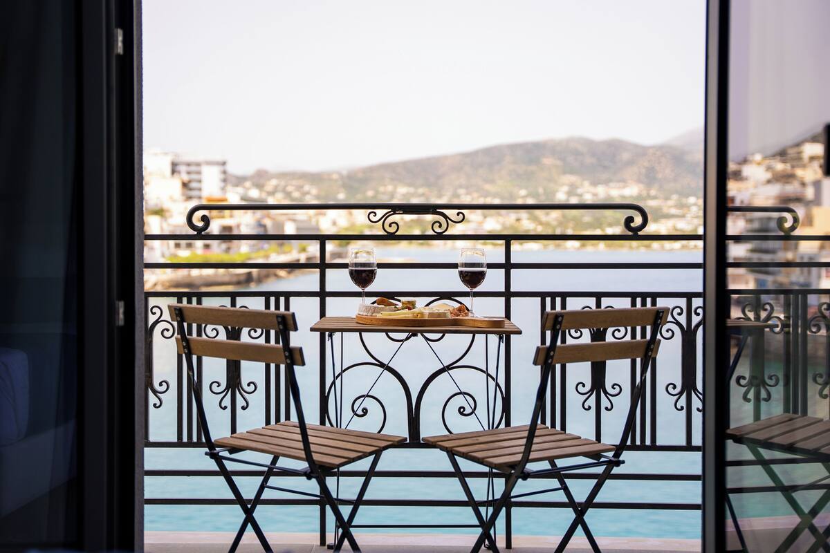 A balcony with two wooden chairs and a table, a railing, and a view of a body of water and distant buildings.