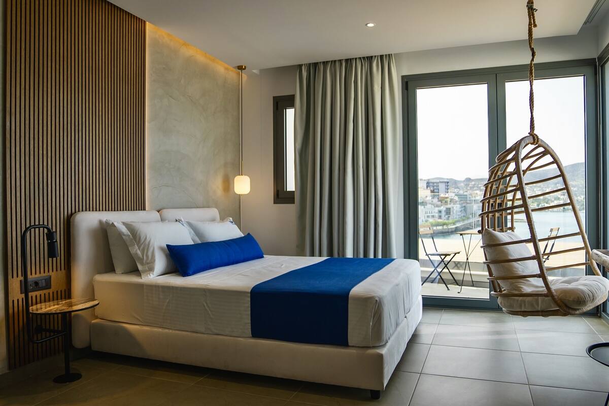 A modern hotel room with a large bed, a hanging chair, a view of the city, and a wooden panel wall.