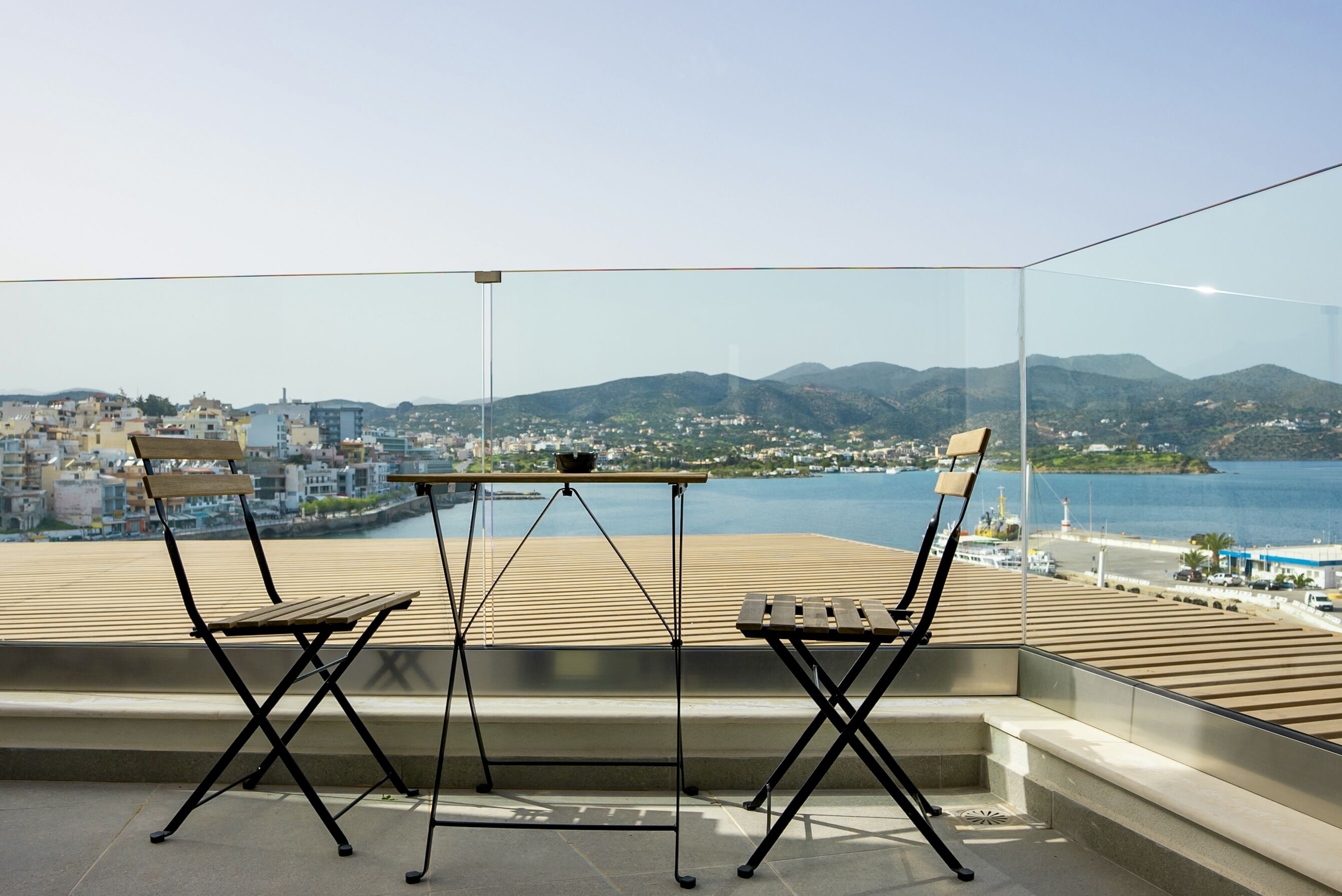 Een balkon met twee houten stoelen en een kleine tafel, met uitzicht op een kustplaats en bergen in de verte.