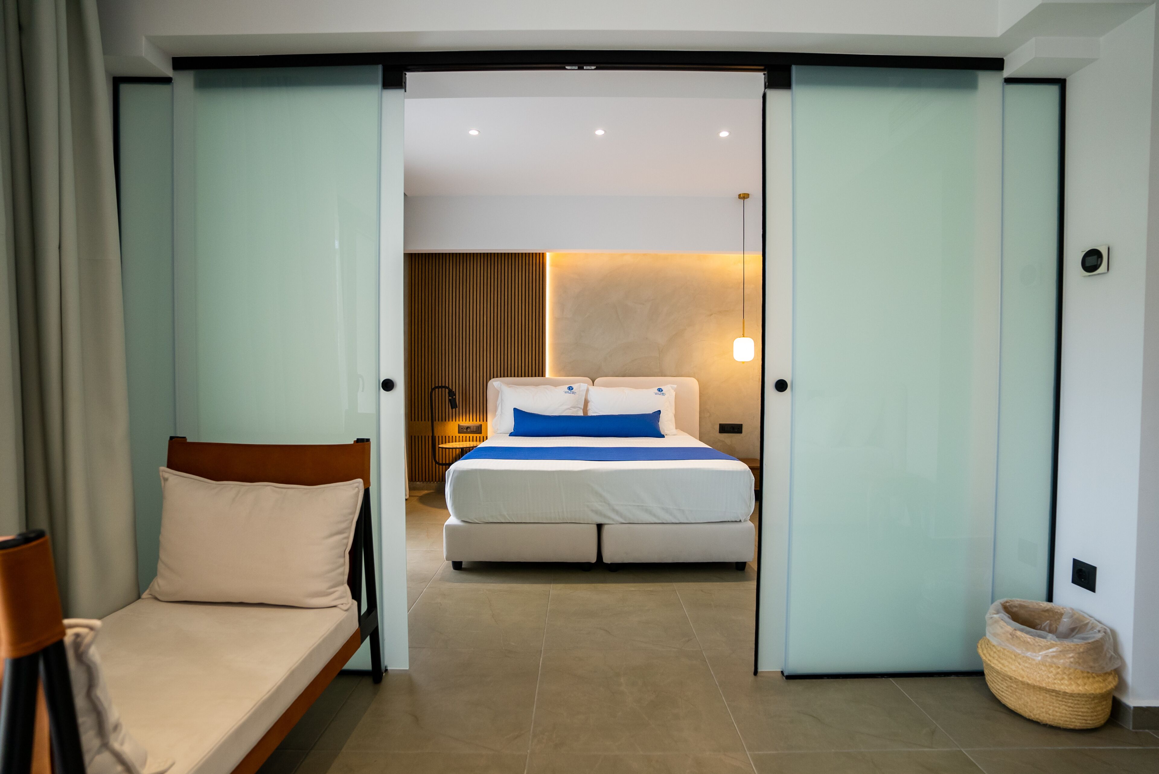 A hotel room with a bed, a small seating area, and a view into another room.