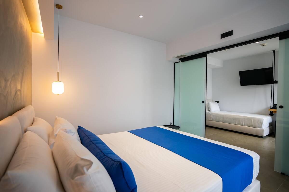 A modern hotel room with a large bed, a bedside table, a flat-screen TV mounted on the wall, and a sliding glass door leading to a balcony.