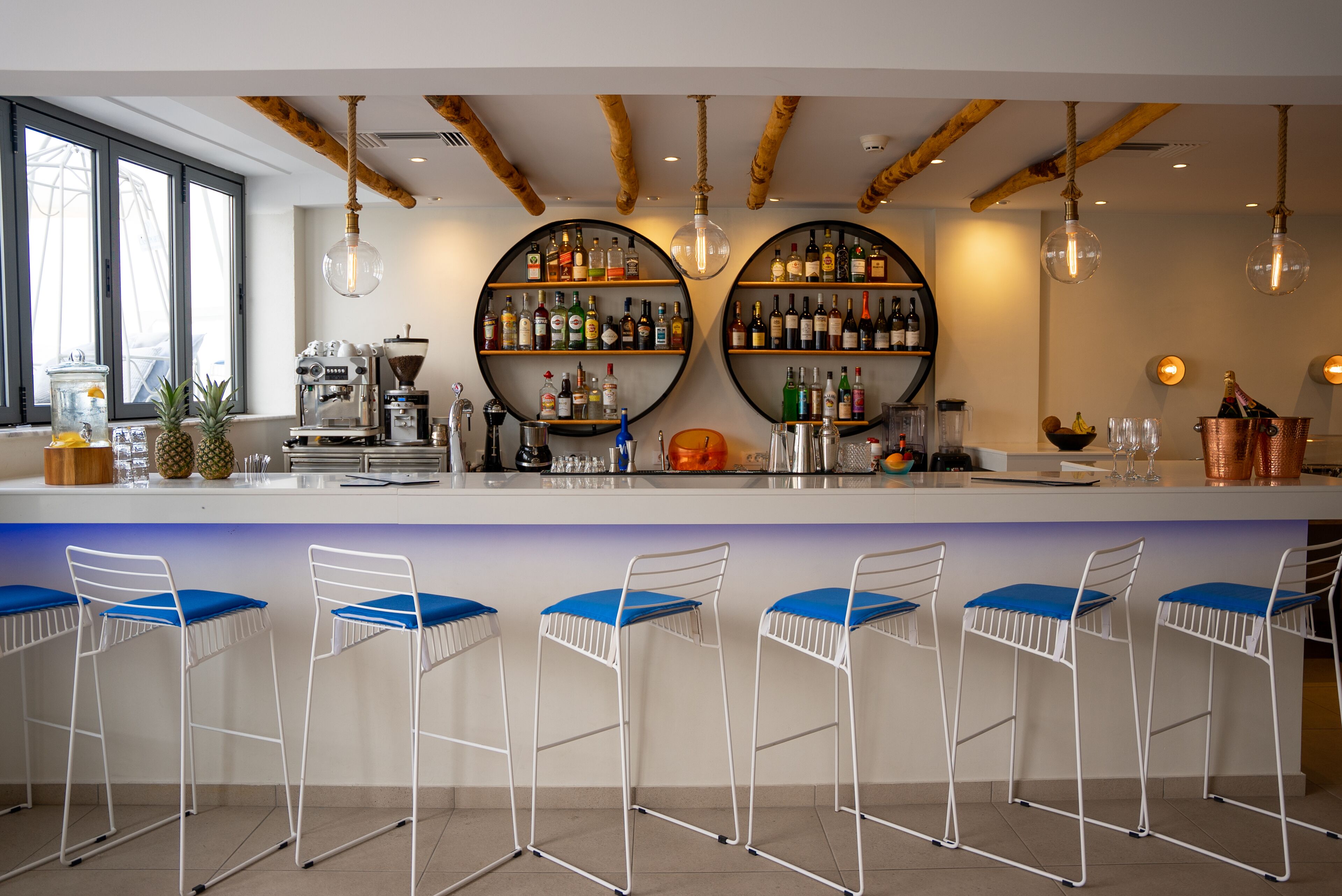 Un bar con una barra blanca, taburetes azules y dos amplias repisas circulares llenas de botellas.