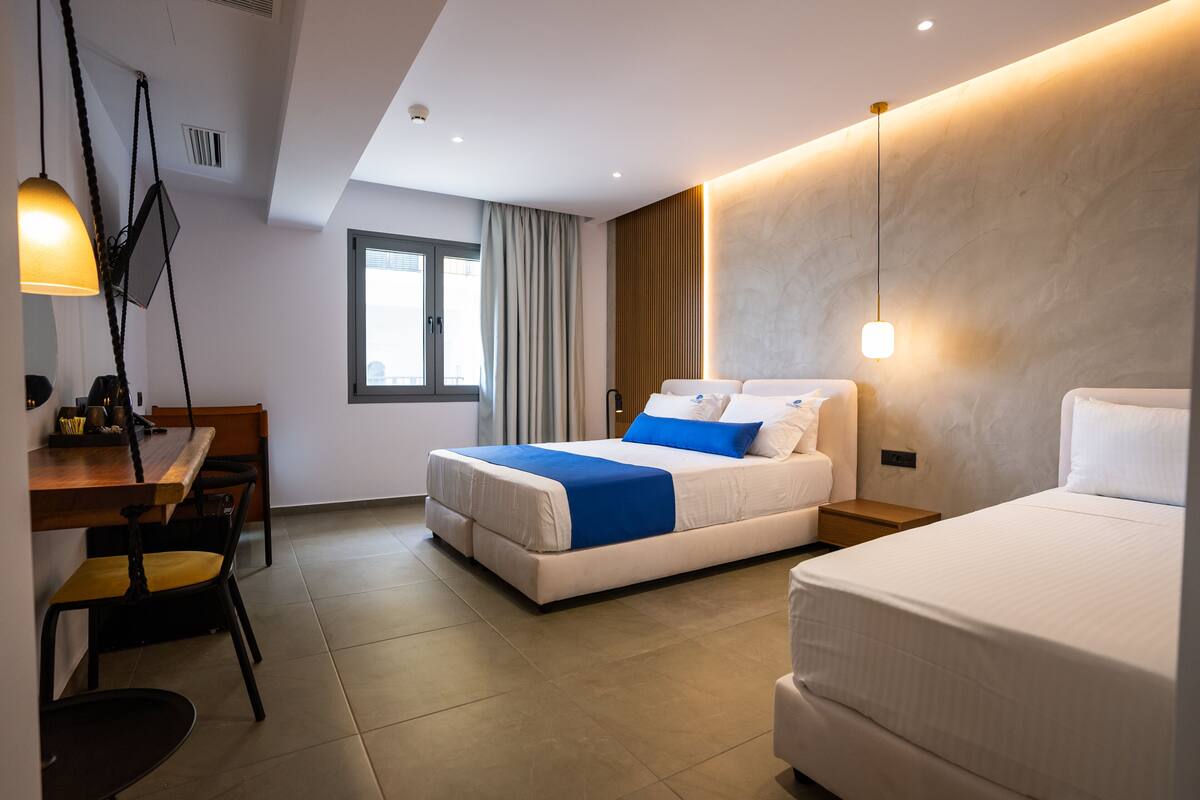 A modern hotel room with a large bed, a smaller bed, a desk with chairs, and a window with curtains.