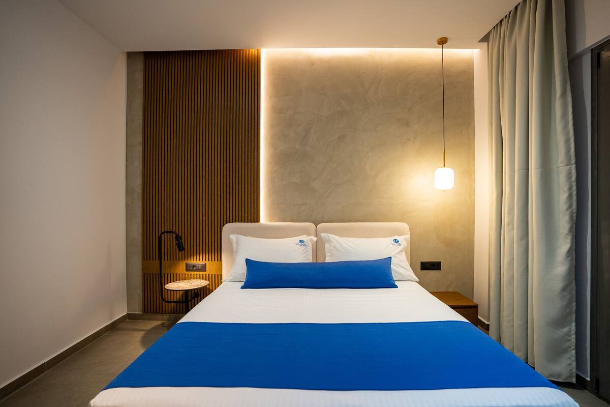 A modern hotel room with a large bed, a bedside table, a wall-mounted light, and a wooden panel design on the left wall.
