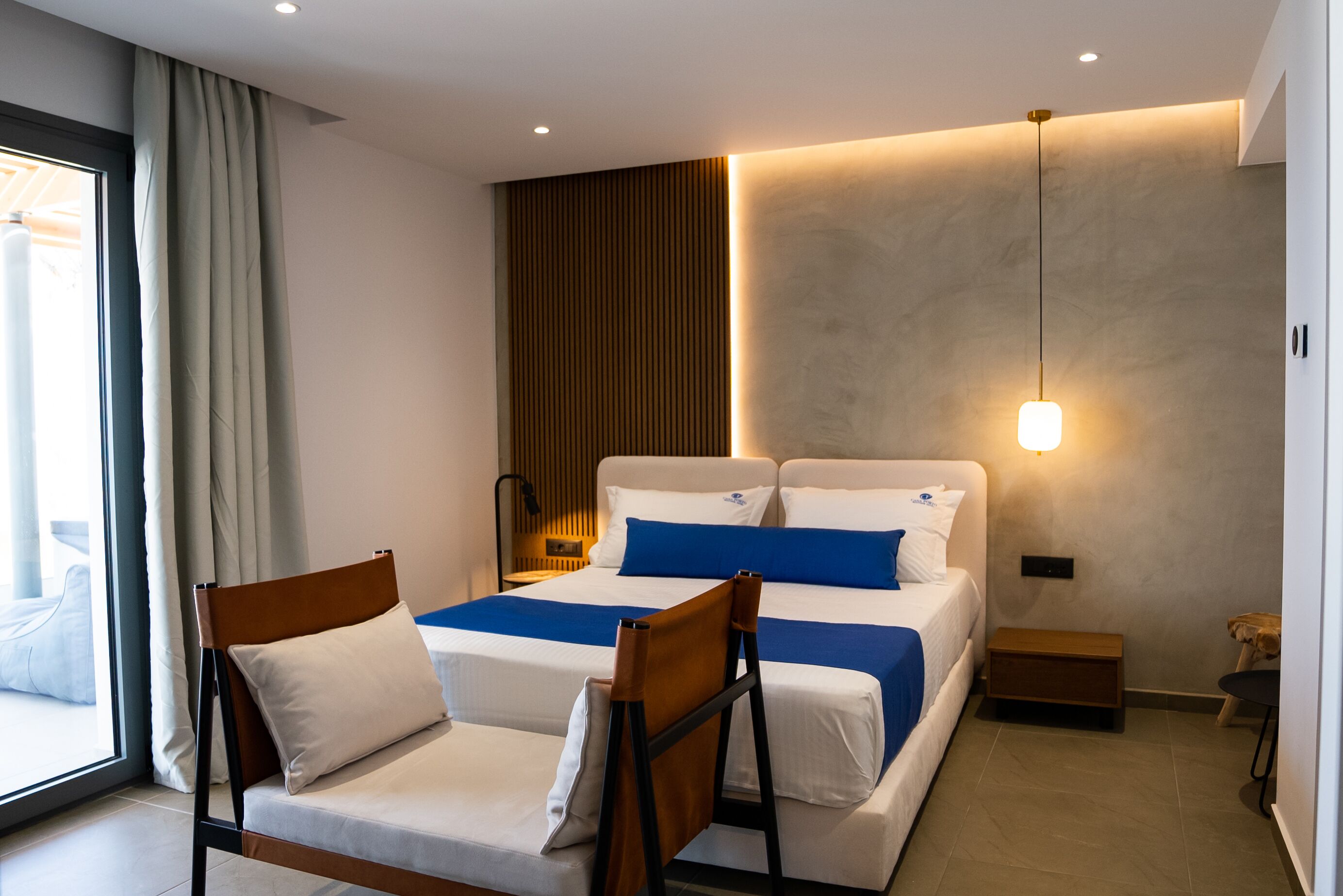 Une chambre d’hôtel moderne avec un grand lit, deux fauteuils, une table de chevet et une vue sur l’extérieur.