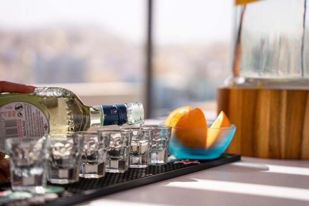 A bottle of liquor being poured into shot glasses on a tray, with a bowl of orange slices and a glass container in the background.