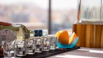 A bottle of liquor being poured into shot glasses on a tray, with a bowl of orange slices and a glass container in the background.