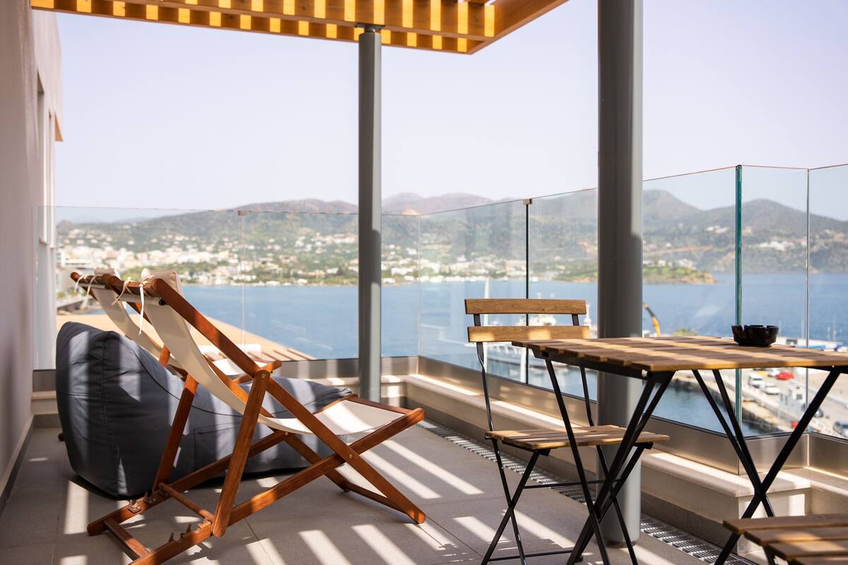 A balcony with a wooden table and chairs, a bean bag, and a view of a coastal town and mountains.