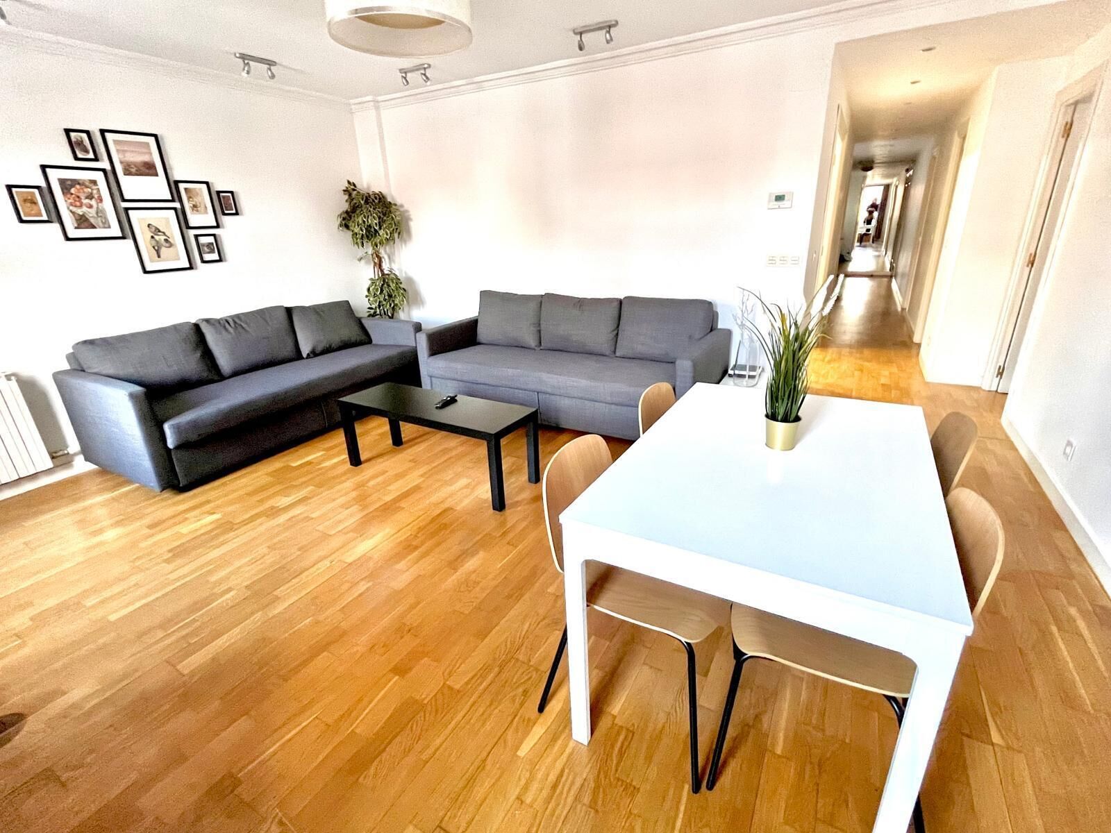 Ein modernes Wohnzimmer mit einem Ecksofa, einem Esstisch mit Stühlen, einem Couchtisch und einem an der Wand hängenden Bilderrahmen.