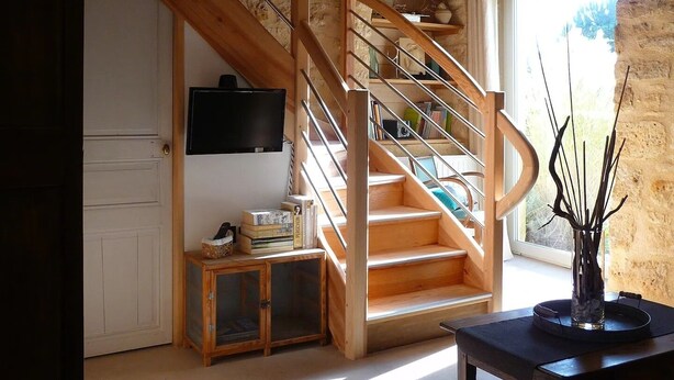 A spacious living area with a wooden staircase, a television mounted on the wall, and a view of the outdoors through a large window.