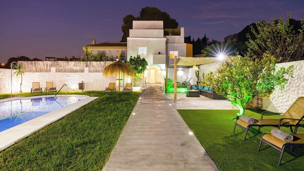 Zona de piscina exterior bien iluminada, con un camino que lleva a un edificio, rodeada de vegetación y sillas de descanso.