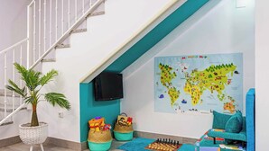 Una escalera con barandilla blanca, un televisor fijado a la pared, un mapa mundial, una planta en una cesta tejida, dos cestas con juguetes y un tablero de ajedrez sobre una alfombra azul.