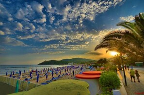 A beach with rows of blue and red lounge chairs, a palm tree, and a sunset in the background. - Hotel Panoramico (Minturno)