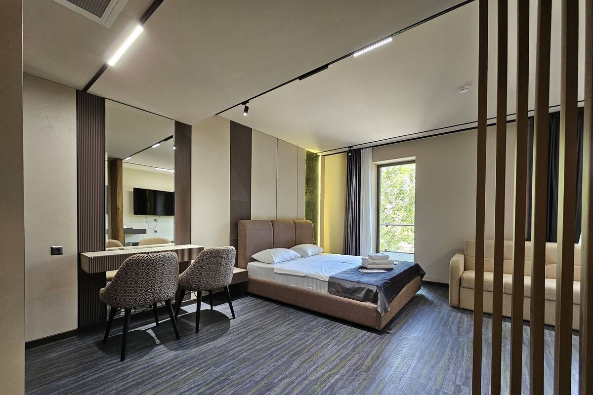 Quarto de hotel moderno com cama, secretária, duas cadeiras estampadas, um espelho grande e vista para o verde lá fora.
